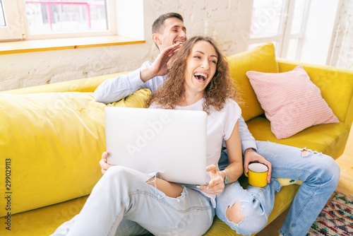 Young happy couple lies on a yellow sofa with a laptop in their apartment, online shopping and internet technology for home