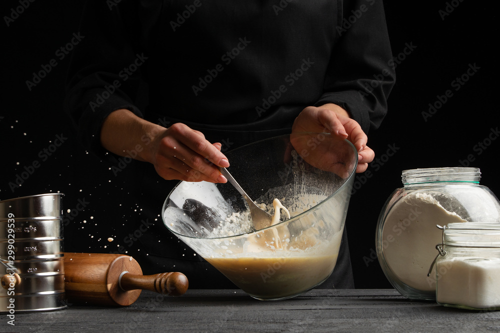 The chef cooks the dough whipping the consistency with a spoon for pasta, pizza, bread and rolls, sprinkles with sugar, freezing in motion. On a black background, horizontal photo - obrazy, fototapety, plakaty 