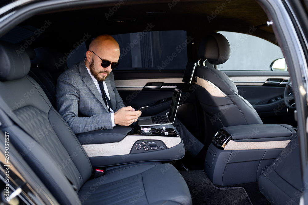 handsome businessman using his mobile phone in a modern car with a driver in center of the city. Concept of business, success, traveling, luxury 