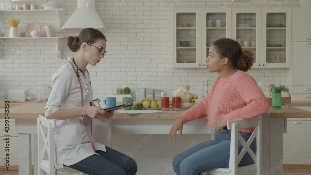 Professional female nutritionist in uniform meeting worried african woman patient in domestic kitchen. Woman dietitian writing diet plan to lovely female patient during medical consultation at home. Professional female nutritionist in uniform meeting worried african woman patient in domestic kitchen. Woman dietitian writing diet plan to lovely female patient during medical consultation at home.