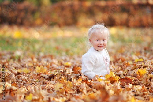 Little cute 3-4-year-old blonde girl sits on fallen leaves and smiles while walking in the autumn park