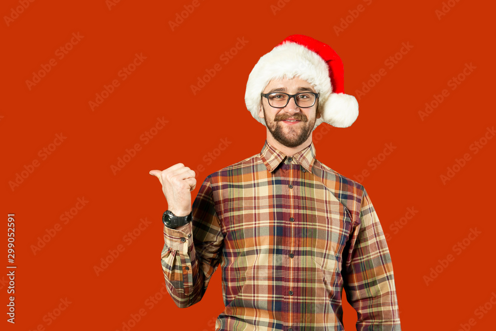 Handsome bearded man in Santa hat and glasses. Santa man pointing to the side over white wall. Christmas concept. Mock up, copy space for text. Hipster bearded man thinking for christmas gift ideas Handsome bearded man in Santa hat and glasses. Santa man pointing to the side over white wall. Christmas concept. Mock up, copy space for text. Hipster bearded man thinking for christmas gift ideas