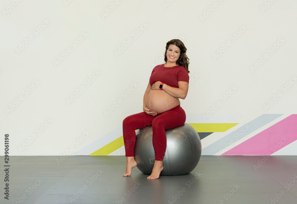Caucasian pregnant woman in her third trimester doing bodyweight floor exercises sitting on a large rubber ball holding her belly in a gym wearing sports or fitness clothing Caucasian pregnant woman in her third trimester doing bodyweight floor exercises sitting on a large rubber ball holding her belly in a gym wearing sports or fitness clothing