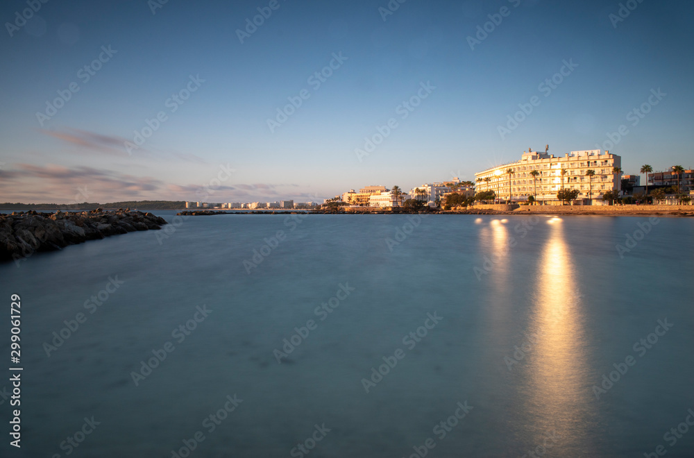 Cala Bona looking towards Cala Millor and the Protur Alicia Hotel at sunrise. Beautiful reflections on the sun in the water which is calm and silky smooth.