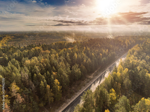 Aerial view on a empty road going through a forest. Sun flare, Nobody, Blue cloudy sky. Fog rises to the sky.