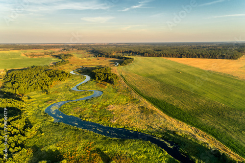 A small river flowing through meadows and agricultural fields. Aerial view. Evening shot with the setting sun.