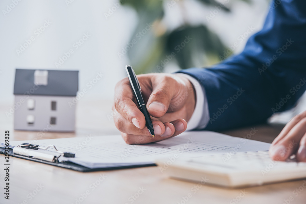 cropped view of businessman using calculator and writing in contract near house model cropped view of businessman using calculator and writing in contract near house model