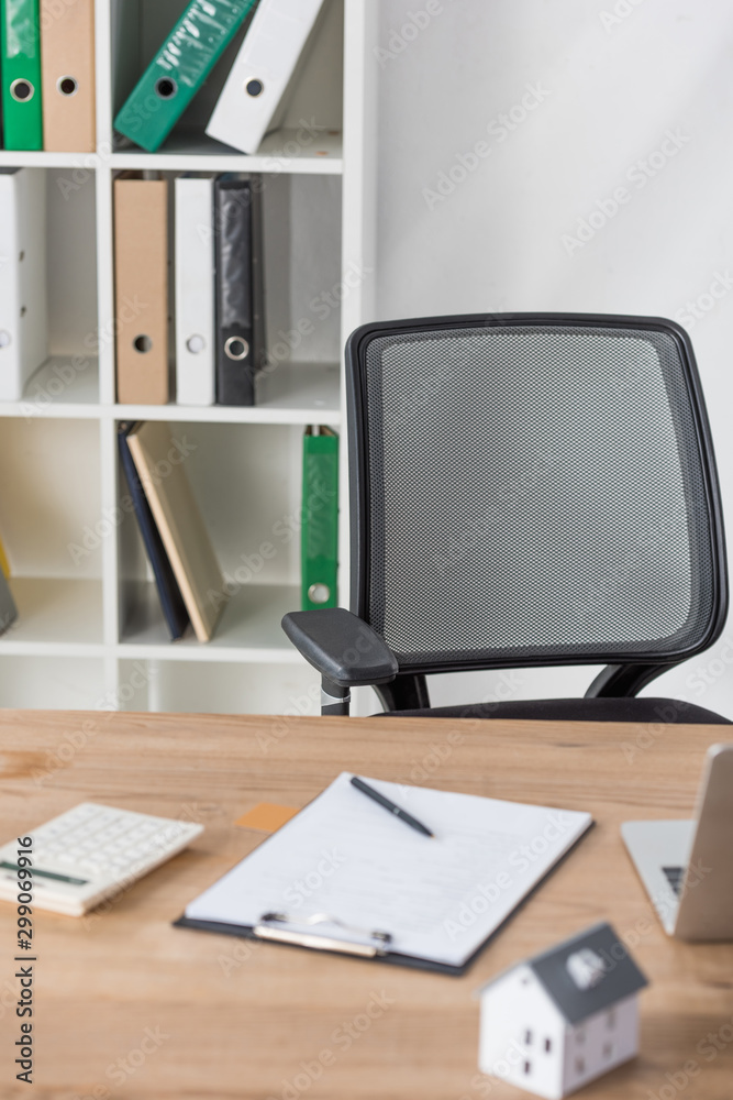 selective focus of office chair and rack with folders near desk with clipboard, calculator and house selective focus of office chair and rack with folders near desk with clipboard, calculator and house