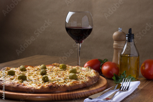 Fototapeta Naklejka Na Ścianę i Meble -  Gratin Loin Pizza. Mozzarella, Canadian Sirloin, Creamy Curd and Green Olives. Accompanied by crostini, glass of red wine, red tomatoes, olive oil, black pepper grinder, fork and knife.