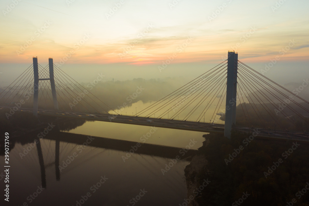 Obraz premium Widok z lotu ptaka na most z samochodami we mgle. Warszawa. Polska. Dron strzelał w ruch pojazdu jadącego w korku na moście nad rzeką. Dron wystrzelił w mgłę o wschodzie słońca nad mostem
