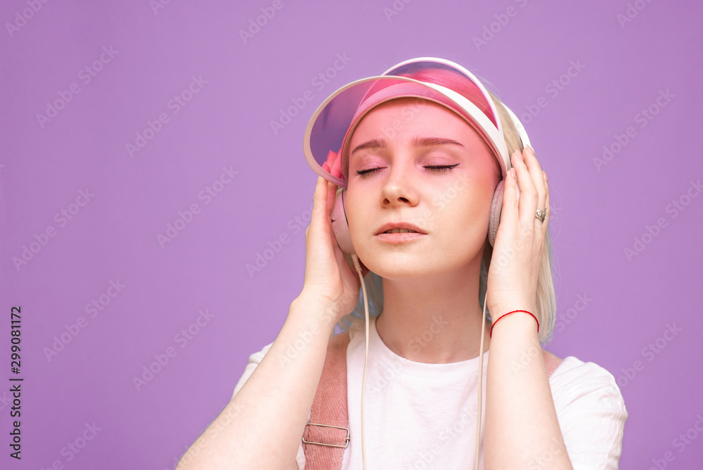 Closeup portrait of a girl in a white T-shirt and a pink cap, listens to music in her headphones with her eyes closed on a purple background. Isolated. Music concept. Closeup portrait of a girl in a white T-shirt and a pink cap, listens to music in her headphones with her eyes closed on a purple background. Isolated. Music concept.