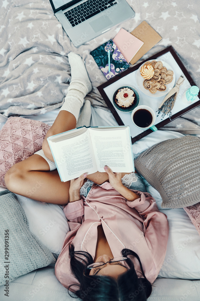 Top view of beautiful young woman in pajamas reading book and enjoying morning coffee while resting in bed at home - obrazy, fototapety, plakaty 