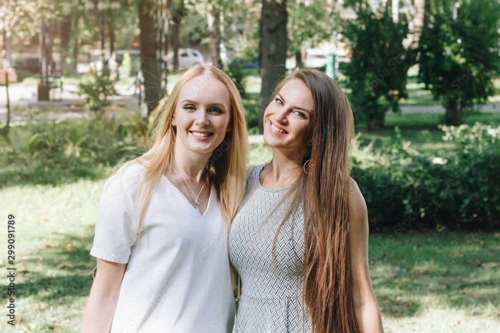 Two beautiful girls brunette and blonde, happy walk in the park, students sisters, girlfriends walk in the sunny summer outdoors. Smiling women, positive emotions Two beautiful girls brunette and blonde, happy walk in the park, students sisters, girlfriends walk in the sunny summer outdoors. Smiling women, positive emotions