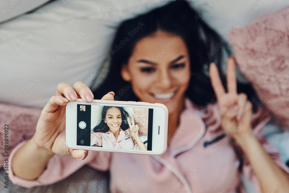Top view of beautiful young woman in pajamas taking selfie and smiling while resting in bed at home Top view of beautiful young woman in pajamas taking selfie and smiling while resting in bed at home