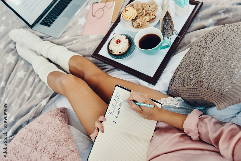 Close up top view of young woman in pajamas writing something down while resting in bed at home - obrazy, fototapety, plakaty 