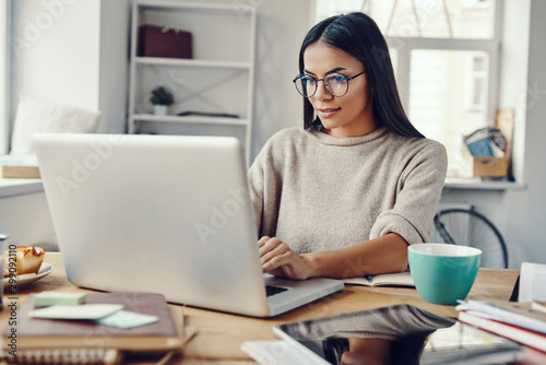 Beautiful young woman in casual clothing using laptop and smiling while working indoors © gstockstudio