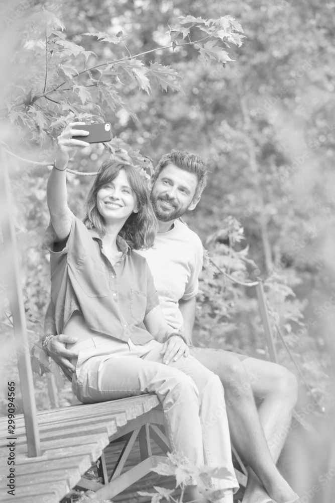 Full length of smiling woman taking selfie with man while sitting on boardwalk at park Full length of smiling woman taking selfie with man while sitting on boardwalk at park