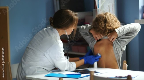 Female doctor and teenager at the clinic. The pediatrician examines the student at the reception. Listens with a stethoscope to the lungs from the back