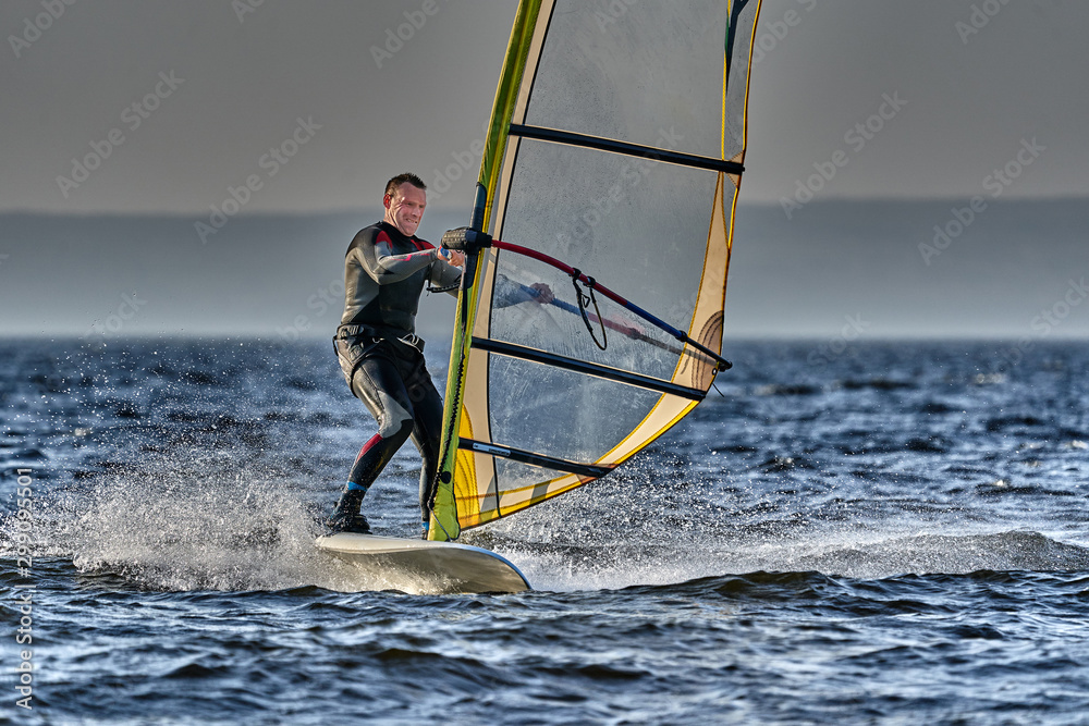 Naklejka premium A male athlete is interested in windsurfing. He moves on a Sailboard on a large lake on an autumn day.