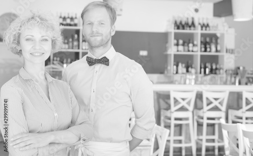 Black and white photo of smiling owner and waiter standing against furniture at restaurant