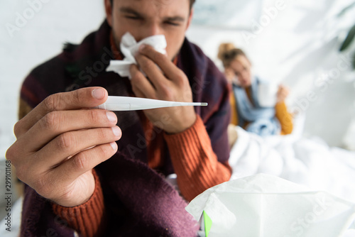 Fototapeta selective focus of sick man with fever holding thermometer and napkin in bed wit