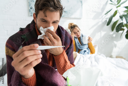 Fotografia sick man with fever holding thermometer and napkin in bedroom with woman behind