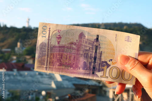 The Reverse Side of Georgian 100 Lari banknote in Woman's Hand with Blurry Tbilisi City View in the Backdrop