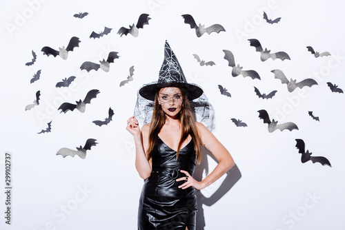 girl in black witch Halloween costume near white wall with decorative bats