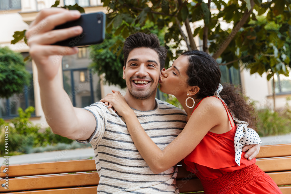Naklejka premium Image of excited young couple taking selfie photo on cellphone and kissing
