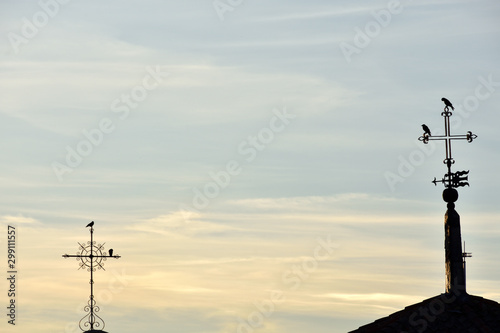 silhouette of two different metal crosses with birds perched on them at sunset and a weather vane pointing to the farthest