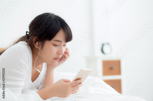 Beautiful young asian woman lying happy earphone listening music online with relax and enjoy in the bedroom, girl fun headphone with playing song smart mobile phone, lifestyle leisure concept.