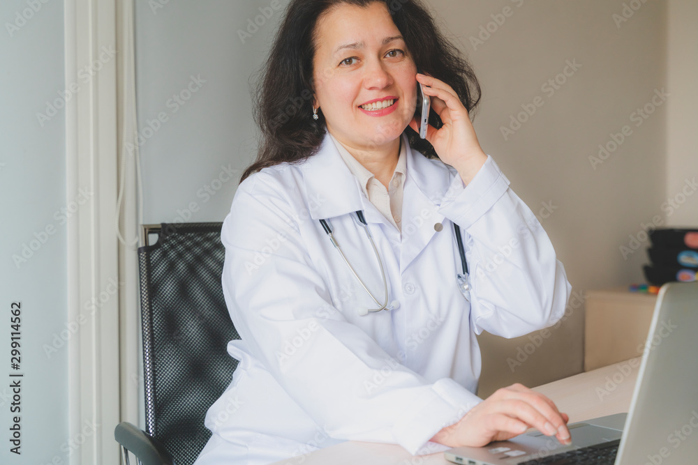 Optomistic doctor woman in medical institution sits at the desktop with computer and speaks on the phone. Phisician is in medical coat with stethoscope Optomistic doctor woman in medical institution sits at the desktop with computer and speaks on the phone. Phisician is in medical coat with stethoscope