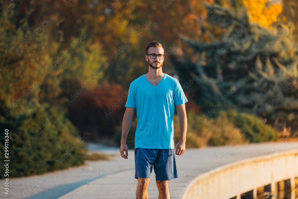 Obraz premium Young man with glasses in sportswear jogging by the lake in a city park. Runner training in the park by the water at sunset.