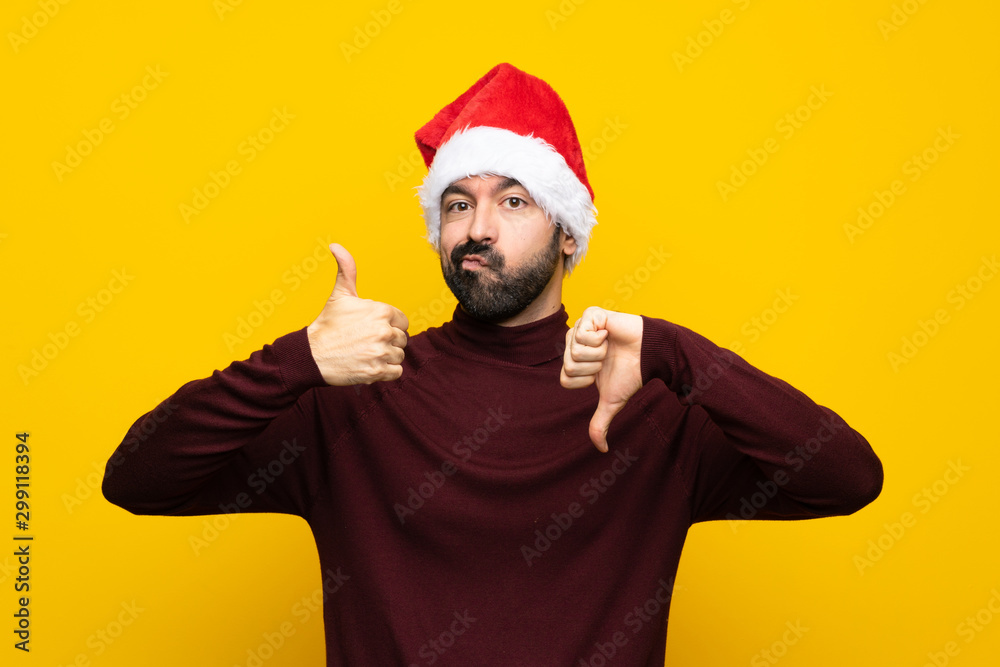 Man with christmas hat over isolated yellow background making good-bad sign. Undecided between yes or not Man with christmas hat over isolated yellow background making good-bad sign. Undecided between yes or not