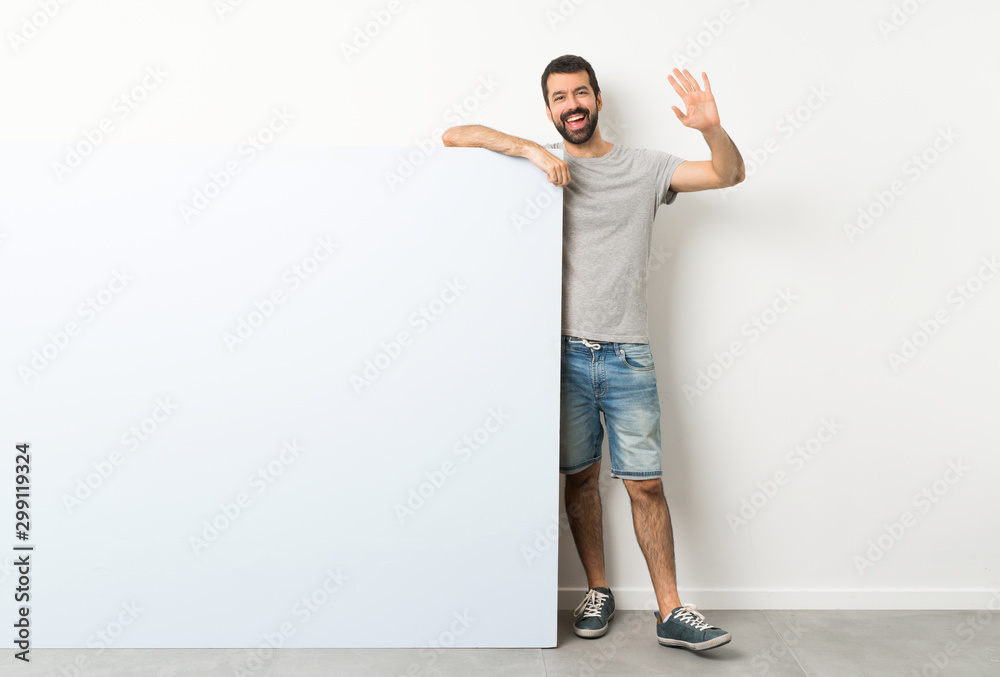 Young handsome man with beard holding a big blue empty placard saluting with hand with happy expression Young handsome man with beard holding a big blue empty placard saluting with hand with happy expression