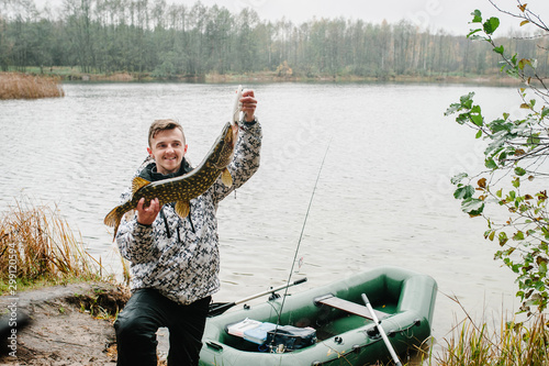 Happy cheerful young fisherman hold a big fish pike near the boat on a background of lake and nature. Fishing background. Good catch. Trophy fish. angler. close up.