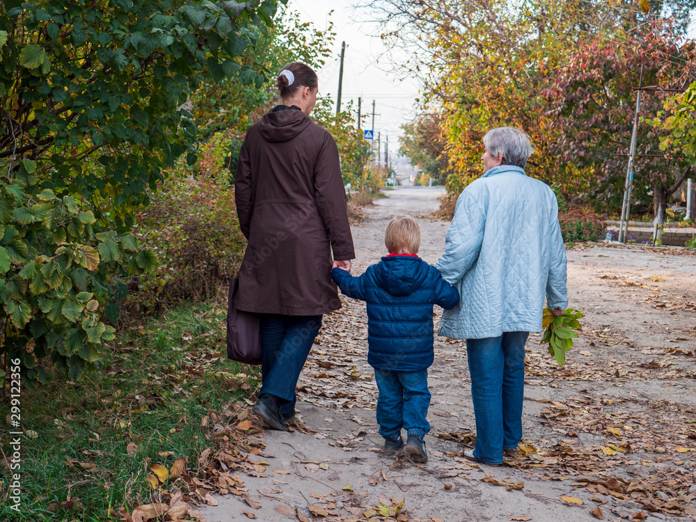 Family on walk. Grandmother, mother and child walk along alley in village. Autumn and fallen leaves Family on walk. Grandmother, mother and child walk along alley in village. Autumn and fallen leaves
