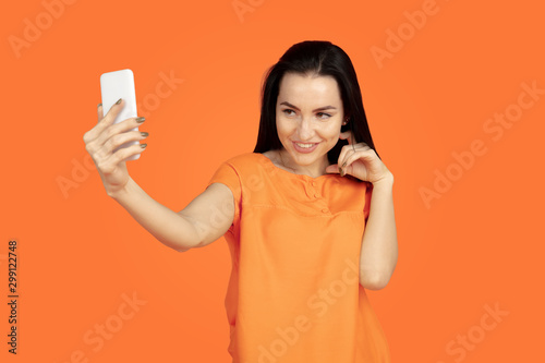 Caucasian young woman's portrait on orange studio background. Beautiful female brunette model in shirt. Concept of human emotions, facial expression, sales, ad. Copyspace. Making selfie, win in bet.