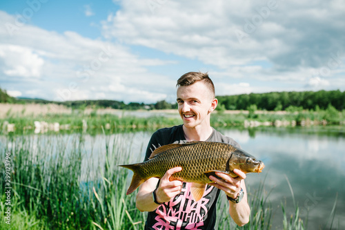 Happy cheerful young fisherman hold a big fish carp on a background of lake and nature. Fishing background. Good catch. Trophy fish. angler.