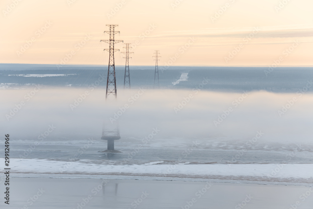 Power lines standing on the Lena River, Yakutia, Republic of Sakha, Russia. Dense fog over the river at sunrise. Beginning of winter. Winter landscape with river and fog. Foggy morning
