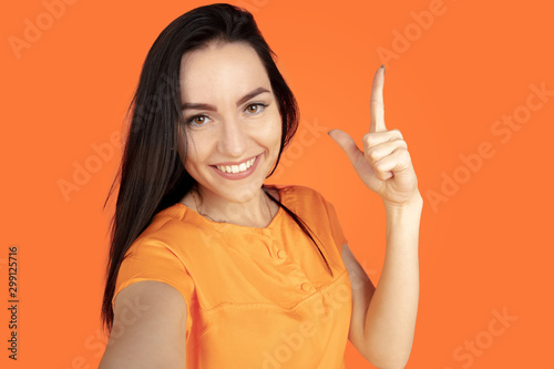 Caucasian young woman's portrait on orange studio background. Beautiful female brunette model in shirt. Concept of human emotions, facial expression, sales, ad. Copyspace. Pointing, showing, smiling.