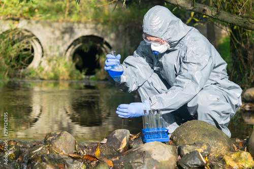 a specialist in a protective suit and respirator analyzes the water in the river near the existing plant, using a flask and a pipette
