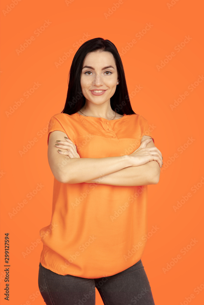 Obraz premium Caucasian young woman's portrait on orange studio background. Beautiful female brunette model in shirt. Concept of human emotions, facial expression, sales, ad. Copyspace. Standing hand crossed.