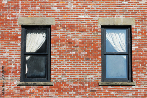 Two windows in a red brick wall. Window ledges are concrete, frames are wooden, painted black. Windows have white curtains, pulled right in one window and left in the other.