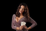 Brunette girl wearing shiny dress posing holding two playing cards in her hand standing against black studio background. Casino, poker. Close-up.