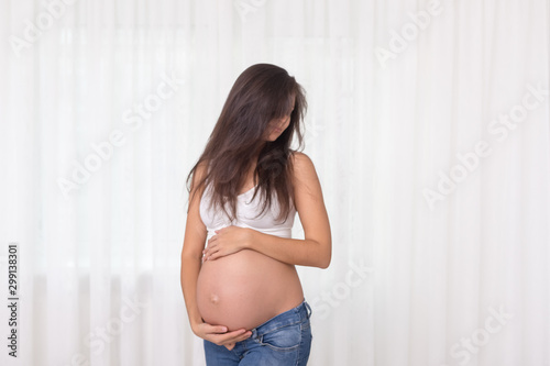Pregnant woman in jeans and white top holds hands on belly on a white background. Pregnancy, maternity, preparation and expectation concept. Beautiful tender mood photo of pregnancy. Free Download Pregnant woman in jeans and white top holds hands on belly on a white background. Pregnancy, maternity, preparation and expectation concept. Beautiful tender mood photo of pregnancy. - GFXLoad