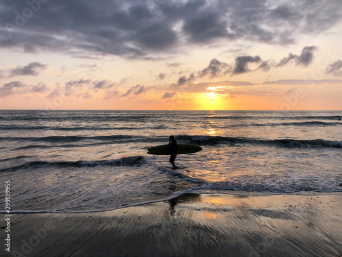 Silhouette of surfer with his board on the beach at Sunset. A man is walking with a surf in his hands across the sea shore during sunset.