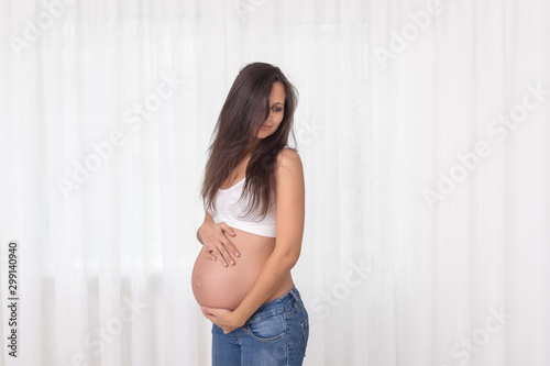 Pregnant woman in jeans and white top holds hands on belly on a white background. Pregnancy, maternity, preparation and expectation concept. Beautiful tender mood photo of pregnancy. Free Download Pregnant woman in jeans and white top holds hands on belly on a white background. Pregnancy, maternity, preparation and expectation concept. Beautiful tender mood photo of pregnancy. - GFXLoad