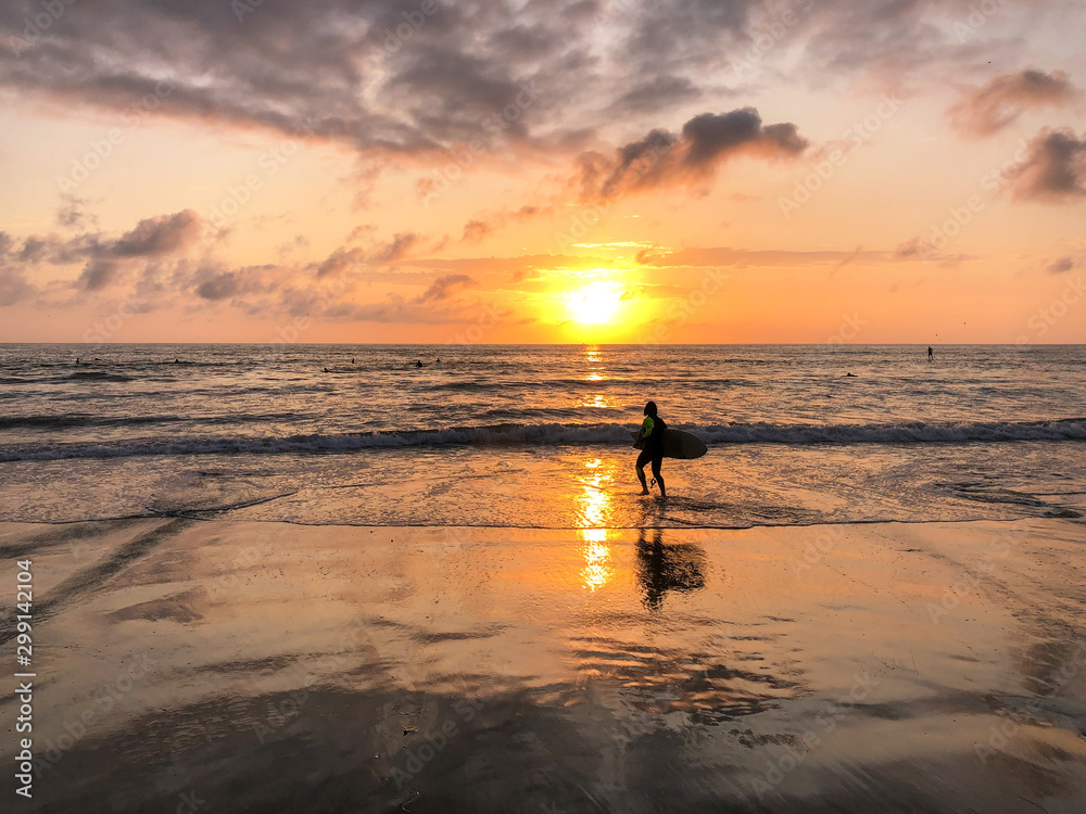 Silhouette of surfer with his board on the beach at Sunset. A man is walking with a surf in his hands across the sea shore during sunset. Silhouette of surfer with his board on the beach at Sunset. A man is walking with a surf in his hands across the sea shore during sunset.