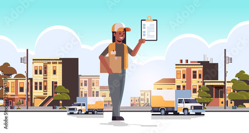 postwoman in uniform holding cardboard parcel box and receiving form african american courier express delivery service concept modern city street cityscape background flat full length horizontal